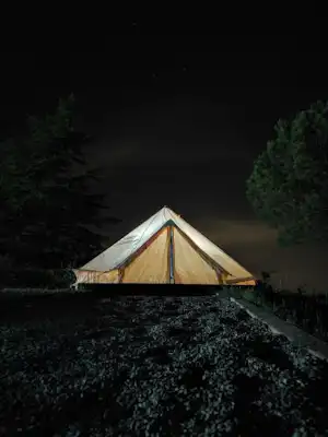 Zelt auf einem Campingplatz bei Nacht, umgeben von Büschen und Bäumen, mit dunklem Himmel im Hintergrund.