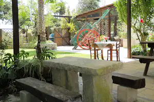 Picknicktisch und Sitzbank im Campinggarten, daneben ein Brunnen aus Stein, umgeben von Bäumen und Pflanzen.