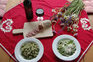 Camping-Frühstück auf roter Decke: Teller mit Kräutergerichten, Holzbrett, Blumenstrauß und Glas mit Getränk.