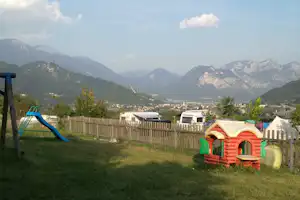 Spielhaus und Rutsche auf einer Wiese am Campingplatz, dahinter Zäune und Wohnwagen mit Blick auf Berge.