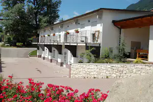 Weiße Ferienhausanlage mit Balkon und Holzgeländer, Steinmauer und Blumenbeeten vor dem Gebäude, gepflasterter Weg und Bäume.