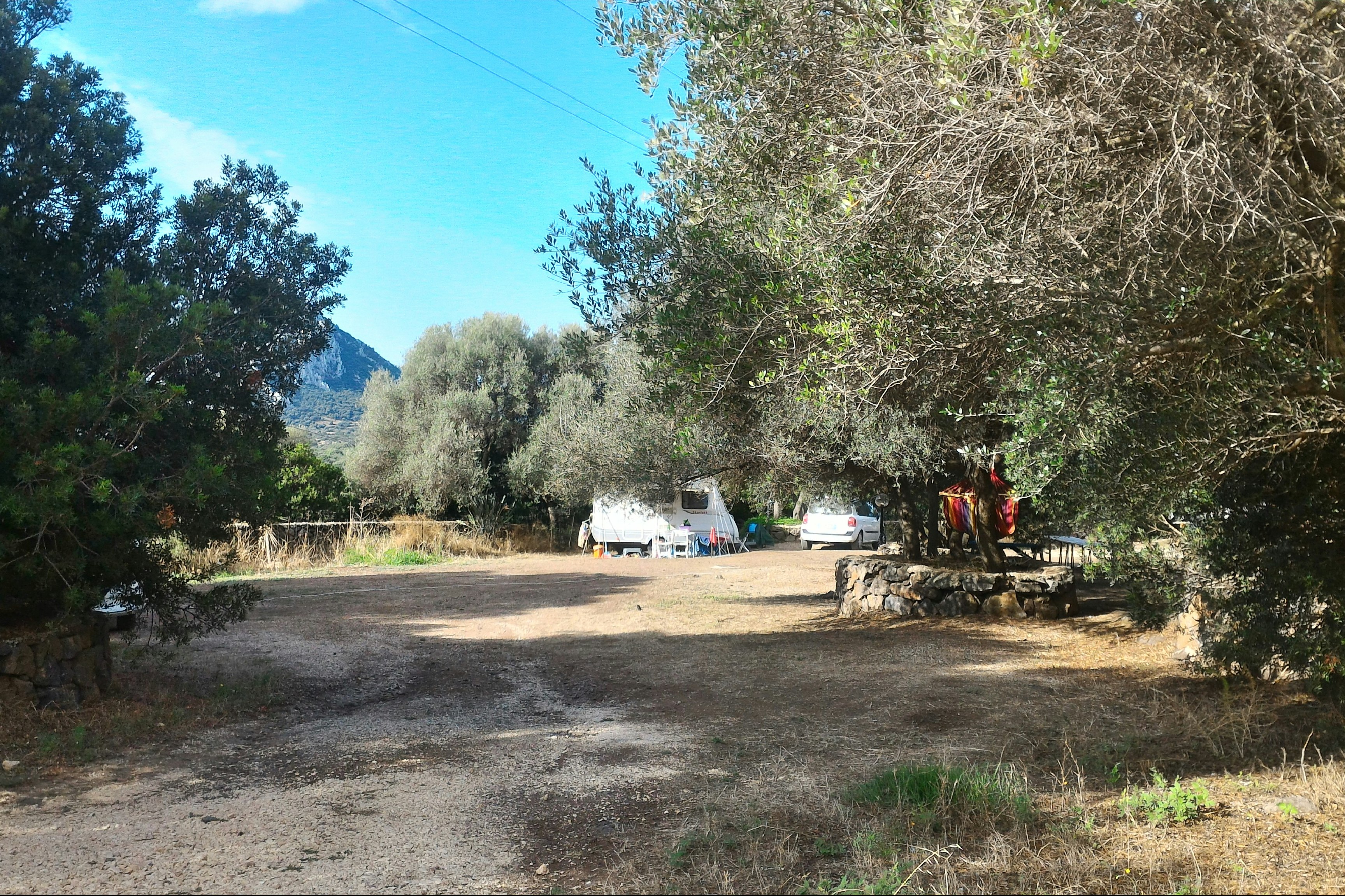Schotterweg im Campingbereich mit mehreren Wohnwagen, Bäumen und Sträuchern am Rand, im Hintergrund ein Berg und blauer Himmel.