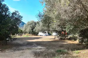 Schotterweg im Campingbereich mit mehreren Wohnwagen, Bäumen und Sträuchern am Rand, im Hintergrund ein Berg und blauer Himmel.