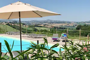 Camping-Pool mit Sonnenschirm, Liegen und Blick auf grüne Hügel und Felder in der Ferne.