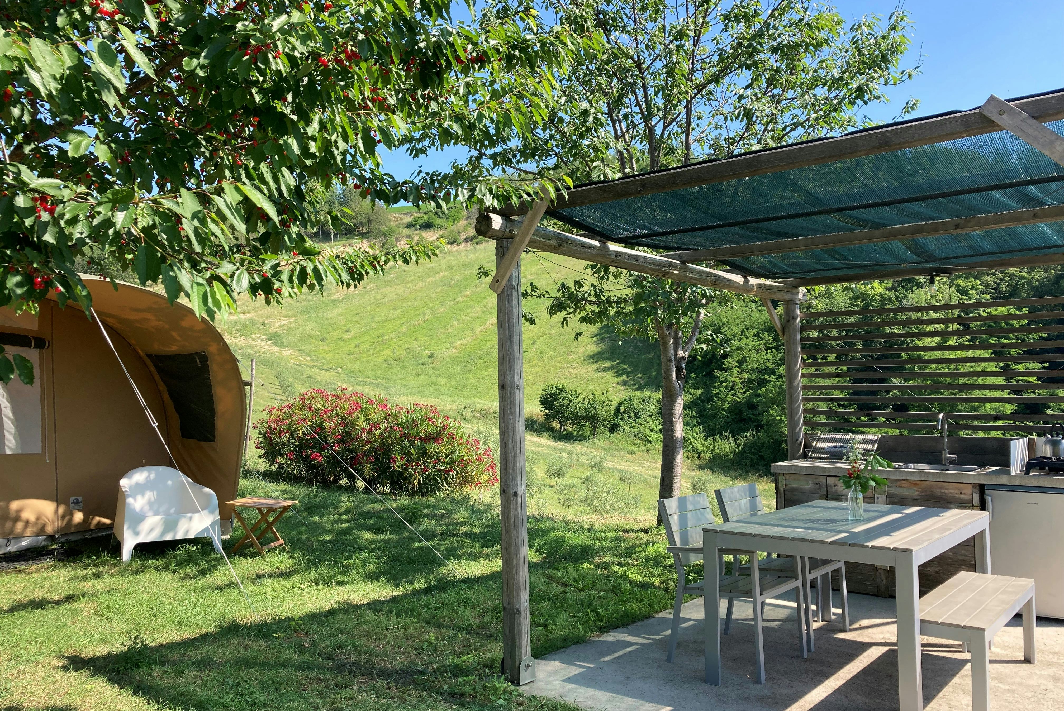 Zelt auf einem Campingplatz-Stellplatz neben einem Pavillon mit Gartenmöbeln, Rasen und Bäumen im Hintergrund.