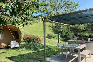 Zelt auf einem Campingplatz-Stellplatz neben einem Pavillon mit Gartenmöbeln, Rasen und Bäumen im Hintergrund.