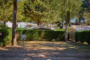 Zelt auf einem Campingplatz zwischen Bäumen, im Hintergrund Wohnwagen und Gebäude, gepflasterter Weg und Stellplatzfläche.