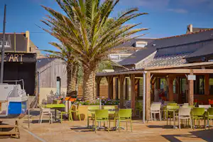 Terrasse mit grünen Stühlen und Tischen, Palmen und Holzdach am Campingplatz, im Hintergrund Gebäude und Wege.