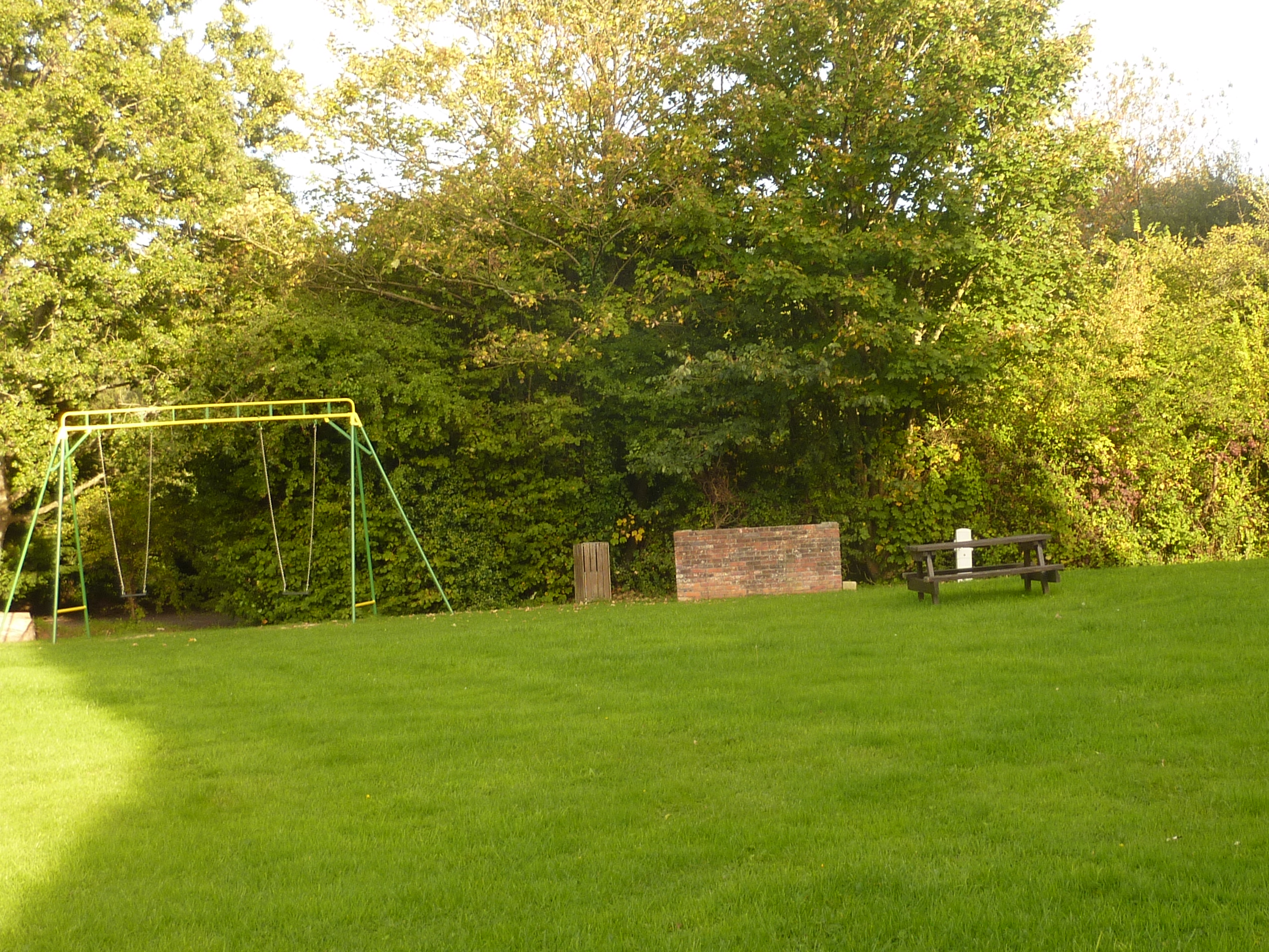 Grüne Campingwiese mit Schaukelgestell, Holzbänken und Spielbereich im Hintergrund, umgeben von Bäumen.