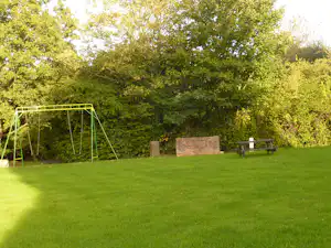Grüne Campingwiese mit Schaukelgestell, Holzbänken und Spielbereich im Hintergrund, umgeben von Bäumen.