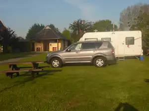 Wohnmobil und silberner SUV auf dem Campingplatz, Rasenfläche mit Bank und Holzhaus im Hintergrund.