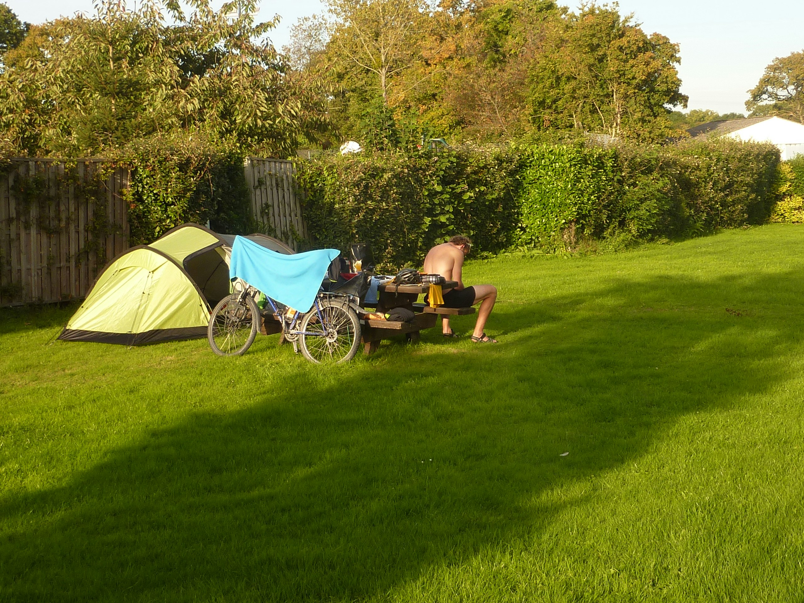 Gelbes Zelt und Fahrrad neben einer kleinen Sitzgruppe auf dem grünen Campingplatz, im Hintergrund Hecken und Bäume.