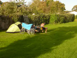 Gelbes Zelt und Fahrrad neben einer kleinen Sitzgruppe auf dem grünen Campingplatz, im Hintergrund Hecken und Bäume.