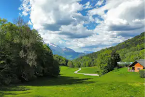 Blick ins Tal mit grüner Wiese, geschwungenem Weg, Campinghütte am Rand und bewaldeten Hängen vor den Alpen.