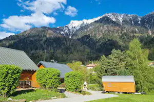 Blick auf die Alpen mit schneebedecktem Bergmassiv, Tannenwald und Campinghäusern mit grünen Büschen im Vordergrund.
