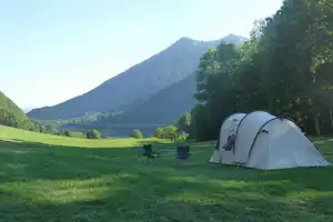 Campingzelt auf einer grünen Wiese, daneben Stühle und Tisch, im Hintergrund Berge und bewaldete Hänge.
