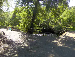 Schotterweg im Campingbereich mit Zaun, Bäumen und Schatten, daneben ein kleiner Bereich mit Wiese.