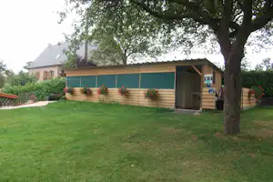 Campinggebäude mit überdachter Terrasse, Holzbänken und Pflanzenkübeln, davor grüner Rasen und Bäume im Hintergrund.
