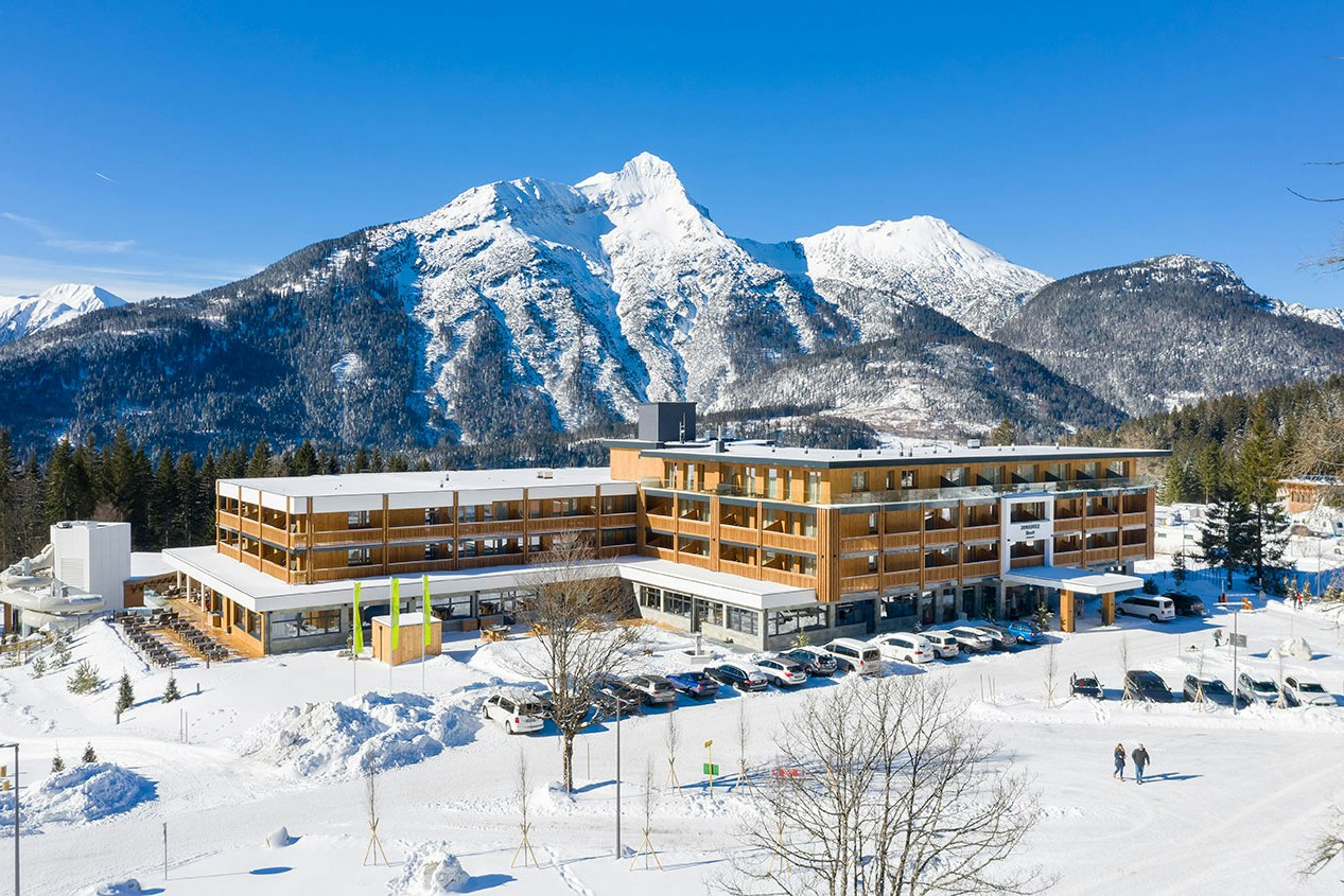 Winterliche Campinganlage mit Holzbungalows, Parkplatz und Wegen im Schnee, umgeben von Bäumen und hohen Bergen.
