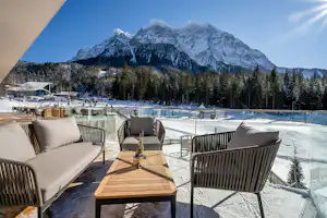 Terrasse mit Holztisch und Sitzgelegenheiten, Blick auf einen Pool und verschneite Berge, umgeben von Nadelbäumen.
