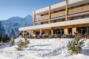 Alpen-Camping-Appartement mit Terrasse, Balkon und Sitzgruppen vor verschneitem Gebäude, im Hintergrund Berge und Tannen.
