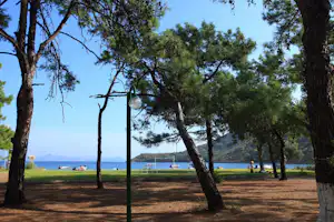 Blick auf einen Campingplatz am See mit Bäumen, Rasenstreifen und Liegewiese am Ufer, im Hintergrund Boote und Berge.