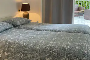 Bett mit gemusterten Bettdecken im Hotelzimmer, daneben Nachttischlampe und Blick durch die Tür auf die Terrasse.