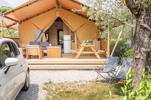 Beiges Zelt mit Holzterrasse, davor ein Stuhl und eine kleine Küchenzeile; links steht ein Auto am Stellplatz.