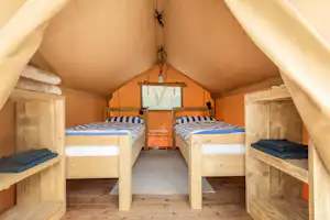 Schlafbereich im Campingzelt mit zwei Betten, Holzrahmen, Deckenleuchte und geöffnetem Fenster im Hintergrund.