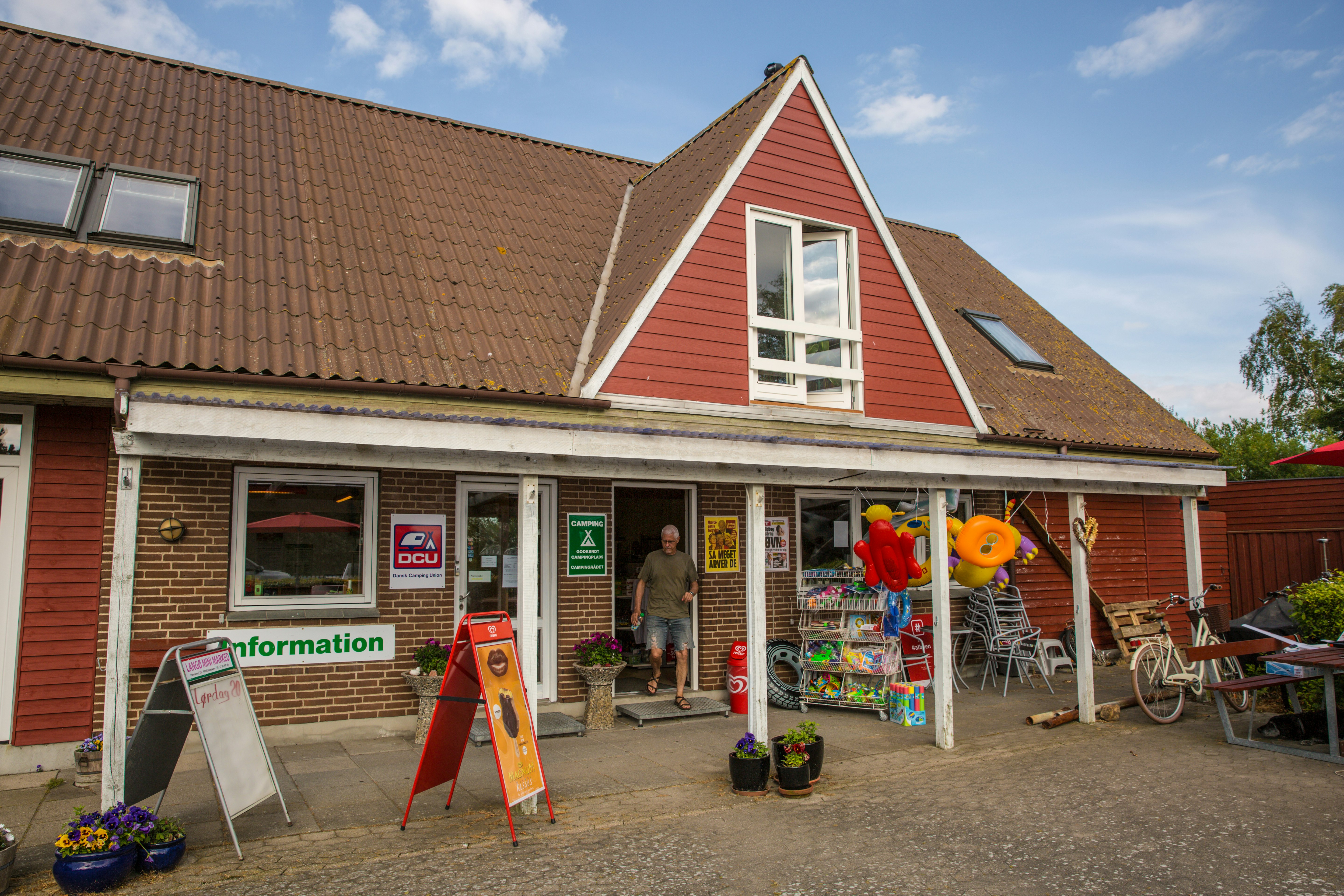 Campinghaus mit rotem Giebel, Eingang und Terrasse, davor Werbetafeln, Fahrräder und Sitzgruppe auf dem Hof.