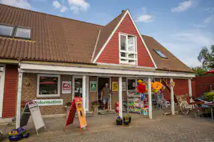 Campinghaus mit rotem Giebel, Eingang und Terrasse, davor Werbetafeln, Fahrräder und Sitzgruppe auf dem Hof.