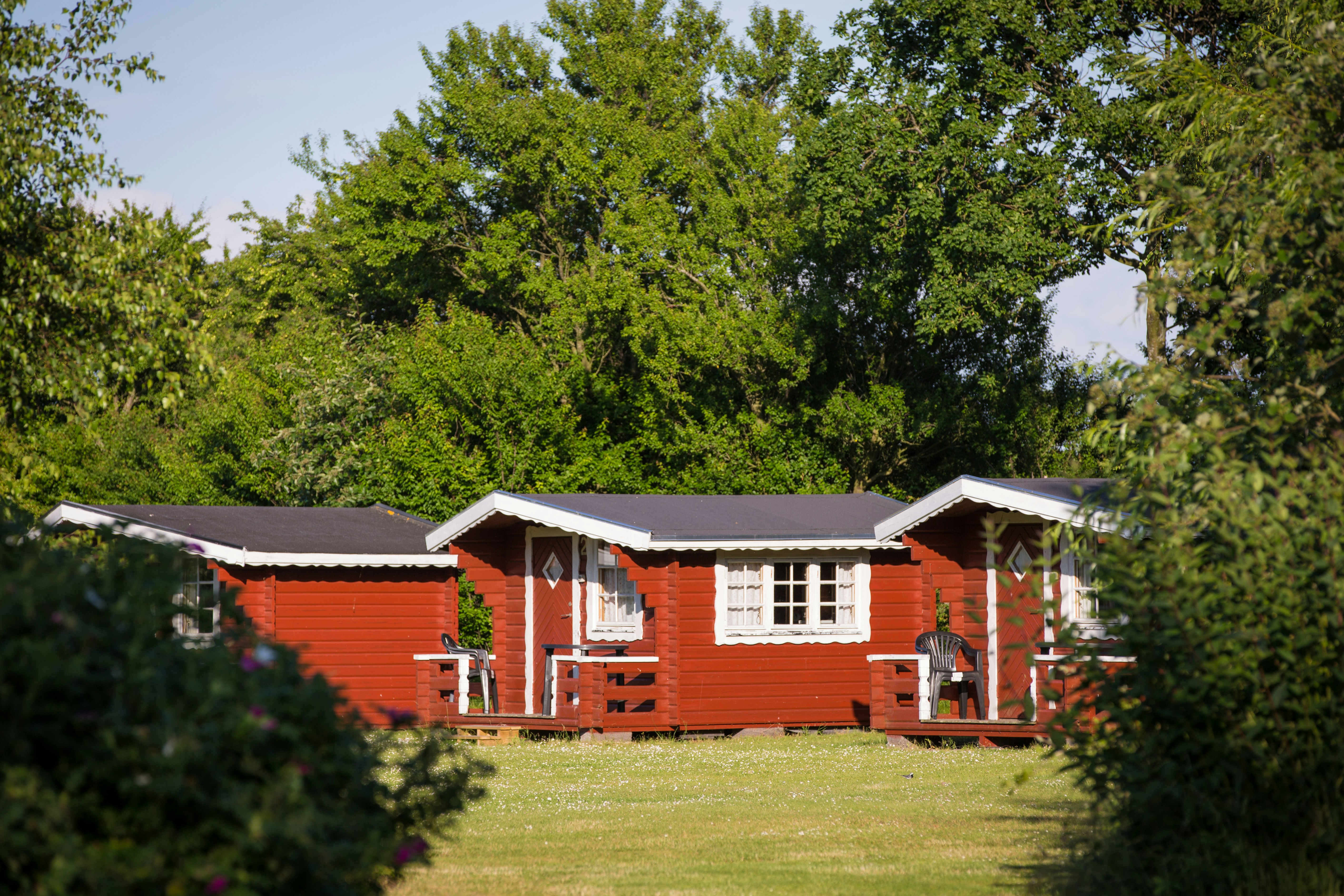 Rotes Ferienhaus mit weißem Fenster und Terrasse auf einer grünen Wiese, umgeben von Bäumen im Campingbereich.