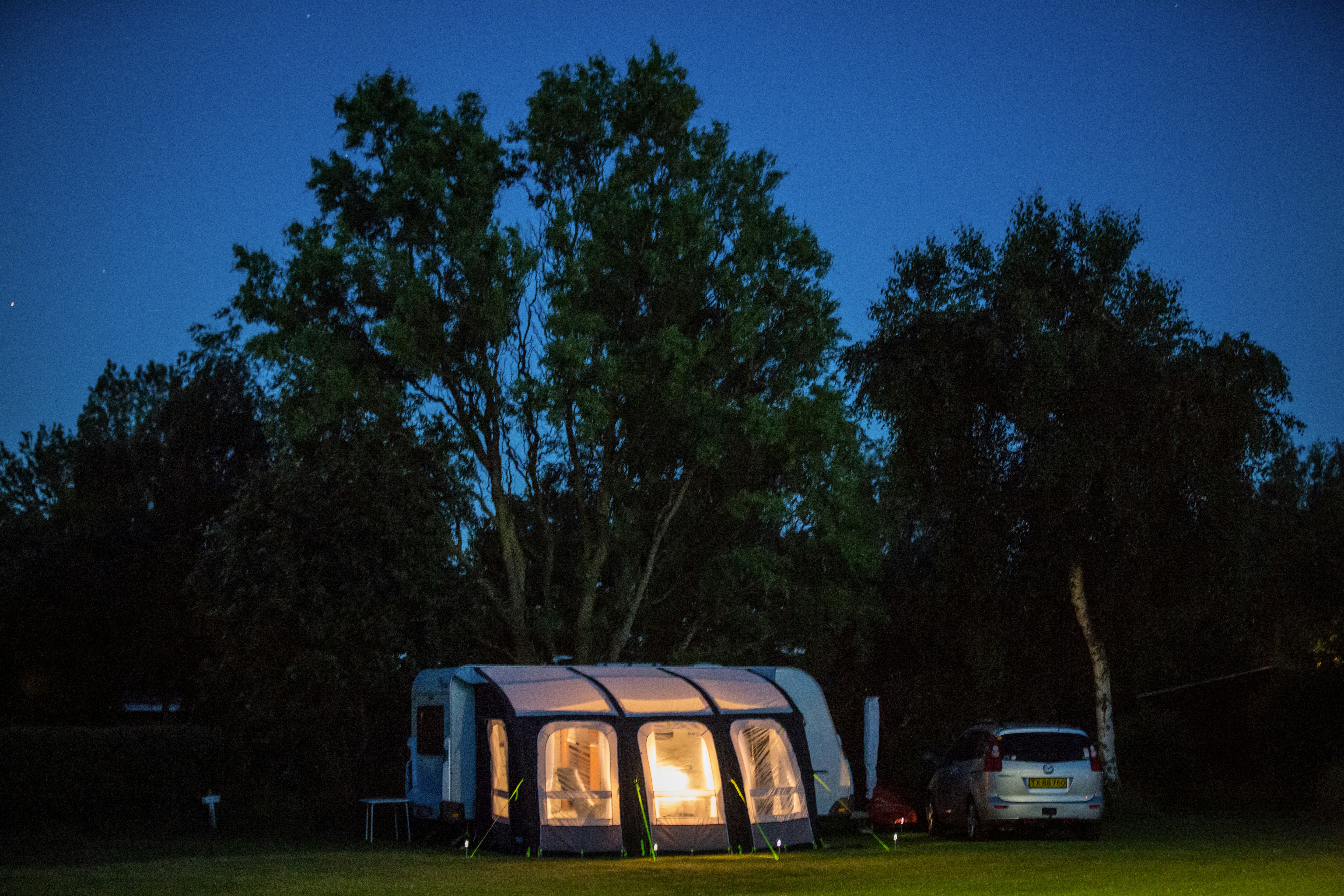 Wohnwagen auf dem Campingplatz im Abendlicht, davor Sitzgelegenheit und Zelt, im Hintergrund Bäume und ein Auto.