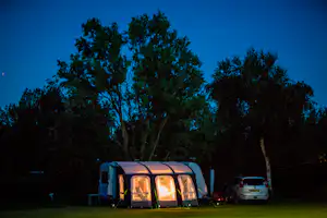 Wohnwagen auf dem Campingplatz im Abendlicht, davor Sitzgelegenheit und Zelt, im Hintergrund Bäume und ein Auto.