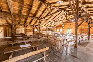 Holzveranda mit überdachtem Bereich, vielen Holzbänken und Tischen, sichtbaren Balken und Stützen im Innenraum.