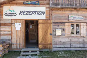 Rezeption eines Campingplatzes in einem Holzgebäude mit Schild „REZEPTION“, Eingangstür und Fenstern.