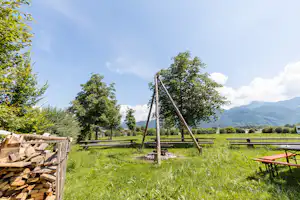 Grüne Campingwiese mit Holzstapeln am Rand, Zaun und zwei Holzpfosten, dahinter Bäume und Berge in der Ferne.