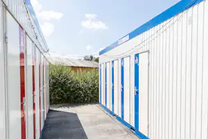 Schmaler Weg zwischen weißen Camping-Containern mit blauen Rahmen, Türen und Hecken im Hintergrund.