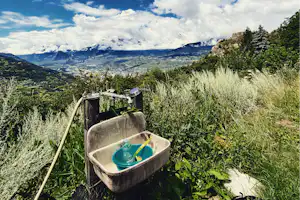 Ein Waschbottich mit blauem Becher auf einer Wiese, dahinter Tal und Berge mit Wolken am Himmel.