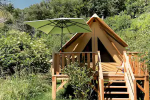 Zeltunterkunft mit Holzveranda und Treppe, Sonnenschirm am Eingang, umgeben von Büschen und Bäumen im Campinggarten.