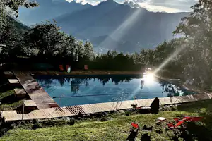 Schwimmbad am Campingplatz mit Holzliegen, Blick auf Berge und Bäume, im Sonnenlicht.
