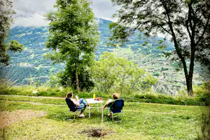 Campingwiese mit zwei Personen am Tisch, umgeben von Bäumen, mit Blick auf grüne Berge im Hintergrund.