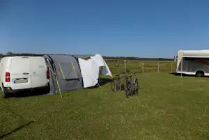 Campingbus mit Vorzelt und Zelt auf einer grünen Wiese, Fahrräder am Stellplatz, im Hintergrund Zaun und Felder.