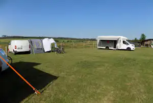 Wohnmobil und Zelt auf einem Campingplatz mit Fahrrädern am Stellplatz, Wiese und Hecke im Hintergrund.