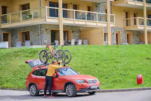 Roter Kleinwagen vor einem Campinggebäude mit Balkonen, davor ein Fahrrad auf dem Dach und grüner Rasen.