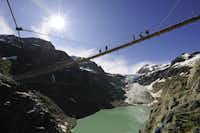 Alpencamping - Brücke mit Wanderern darauf in den Alpen über einem Bergsee