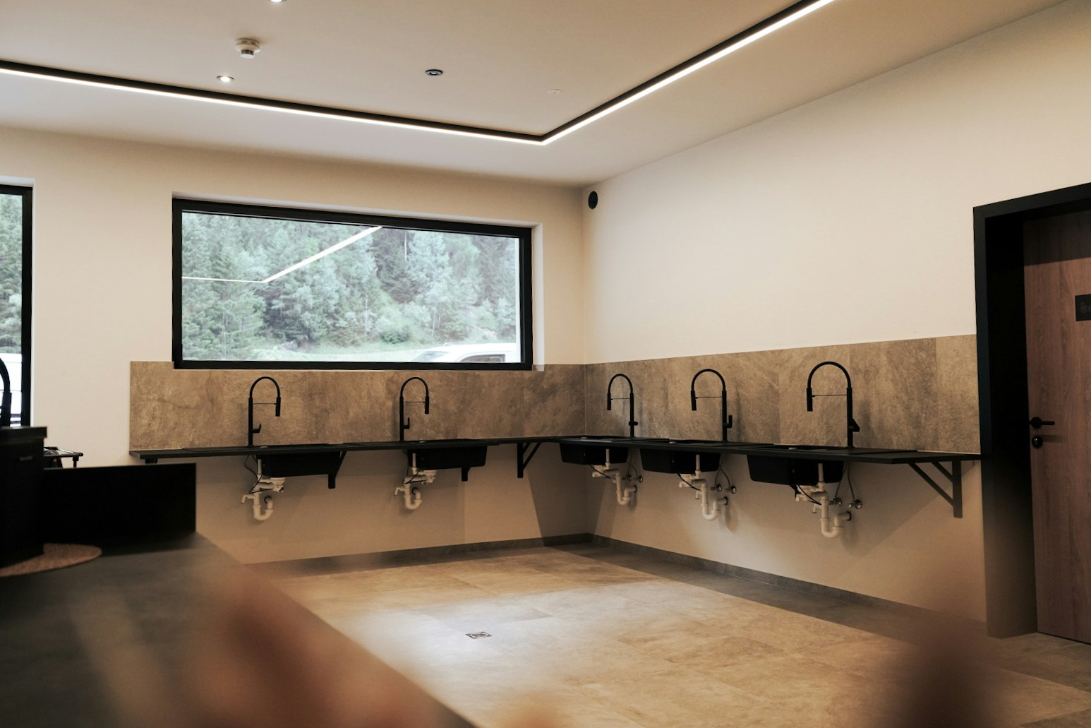 Innenraum mit Betonboden und beleuchteter Decke, an der Wand mehrere Waschbecken mit Hähnen und Spiegeln.