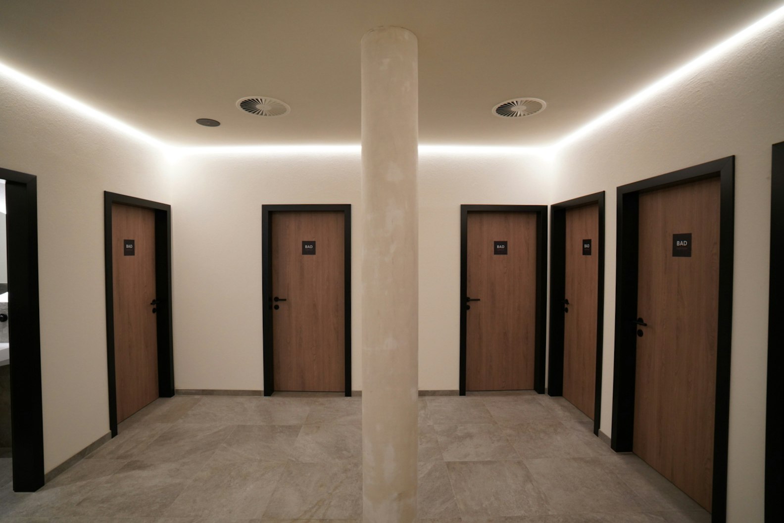 Flur mit mehreren Holztüren, grauem Boden und weißen Wänden, mittig eine Säule mit Deckenbeleuchtung.