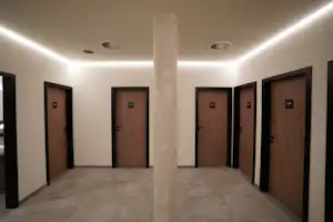 Flur mit mehreren Holztüren, grauem Boden und weißen Wänden, mittig eine Säule mit Deckenbeleuchtung.