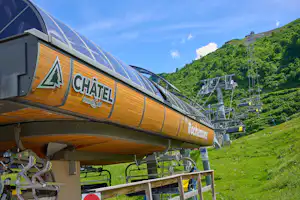 Chalet-Zelt mit Holzverkleidung und Beschriftung, davor Sitzbank und Tisch, im Hintergrund Bergwiese und Seilbahn.